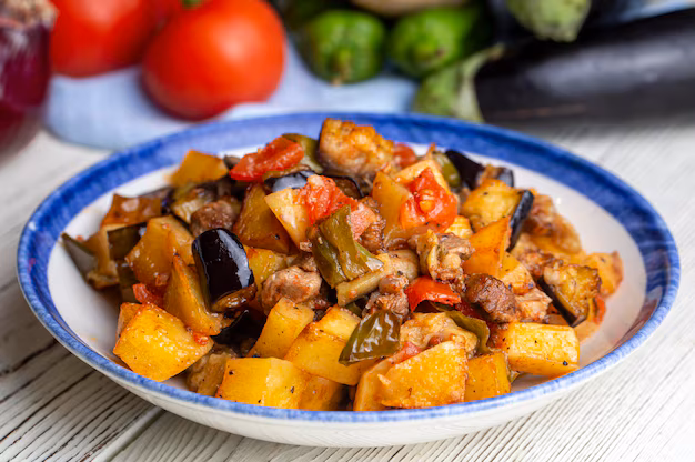 一道豐富的家常法式普羅旺斯燉菜Ratatouille，內含茄子、番茄、甜椒與肉塊，燉煮入味後盛裝於藍邊白色陶瓷盤中。背景擺放著新鮮的番茄、青椒與茄子，整體色澤誘人，呈現出溫暖的地中海家庭料理風格